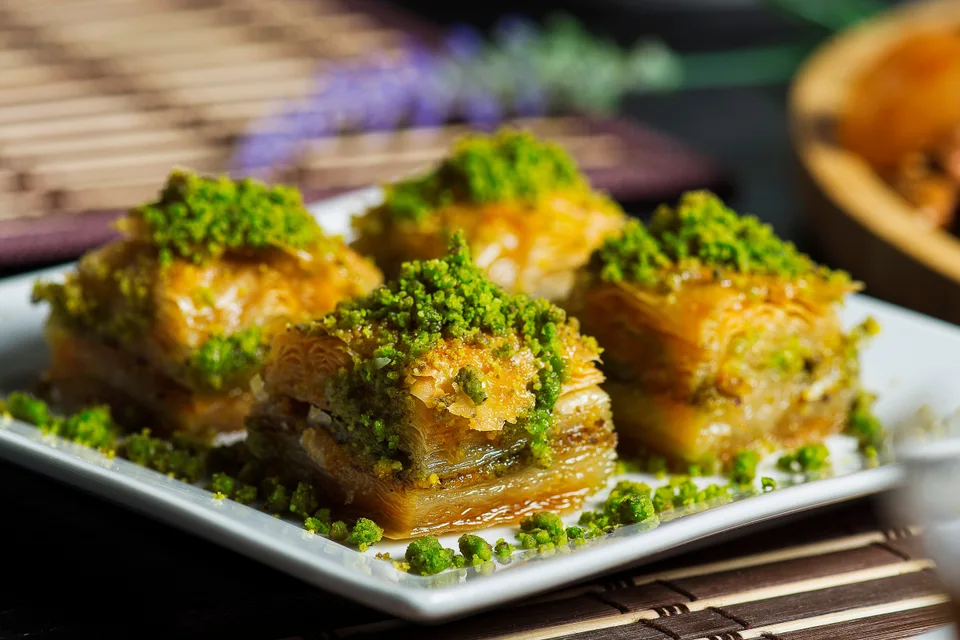 BookNoFees: Beispielfoto: Vier Stück geschichtetes Baklava, garniert mit zerkleinerten Pistazien, auf einem weissen Teller
